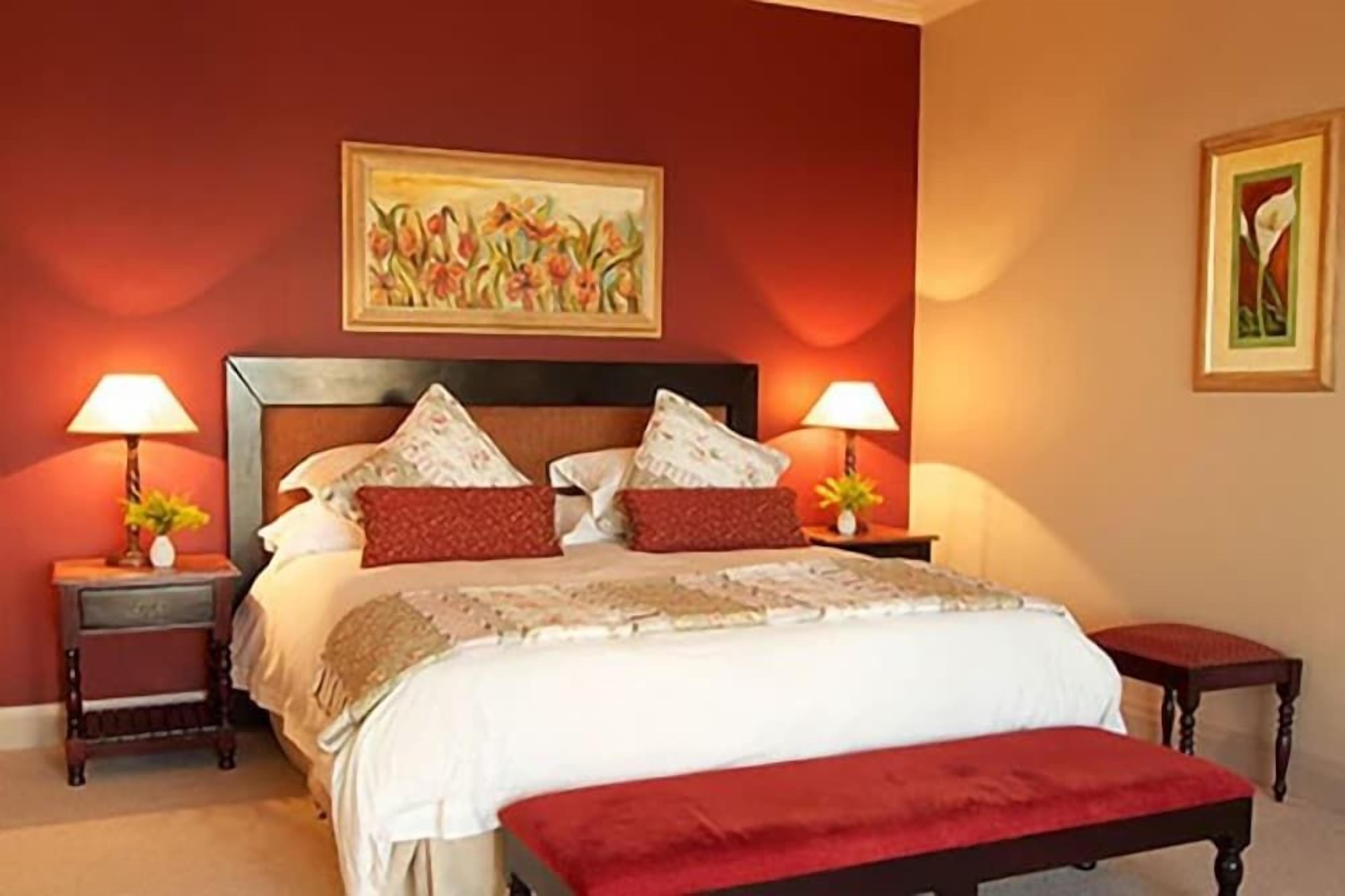 A bedroom with a large bed, two bedside tables with lamps, a bench, and two wall-mounted paintings.