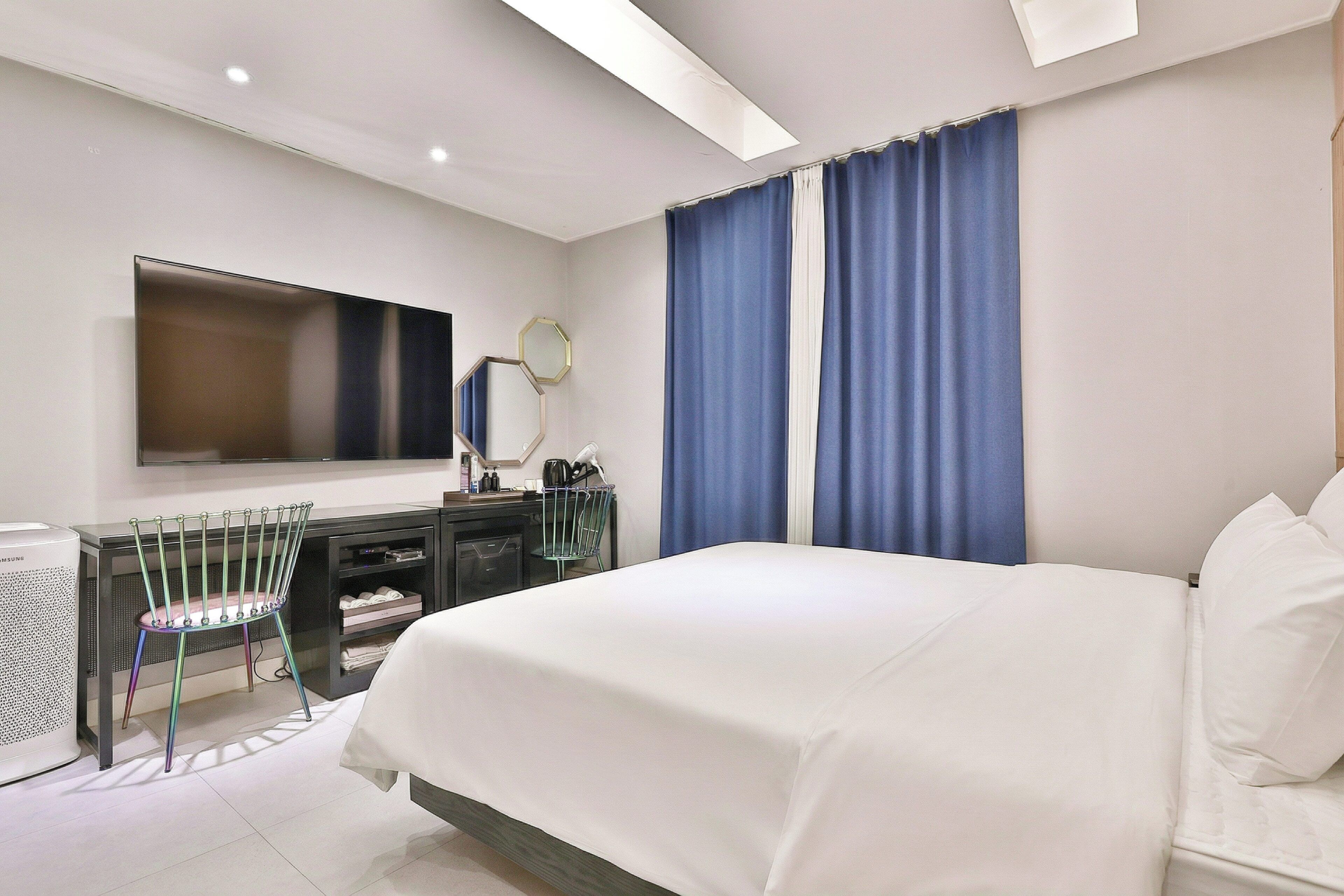 Quarto de hotel moderno com uma cama grande, televisão de tela plana na parede, escrivaninha com cadeira e uma janela ampla com cortinas azuis.