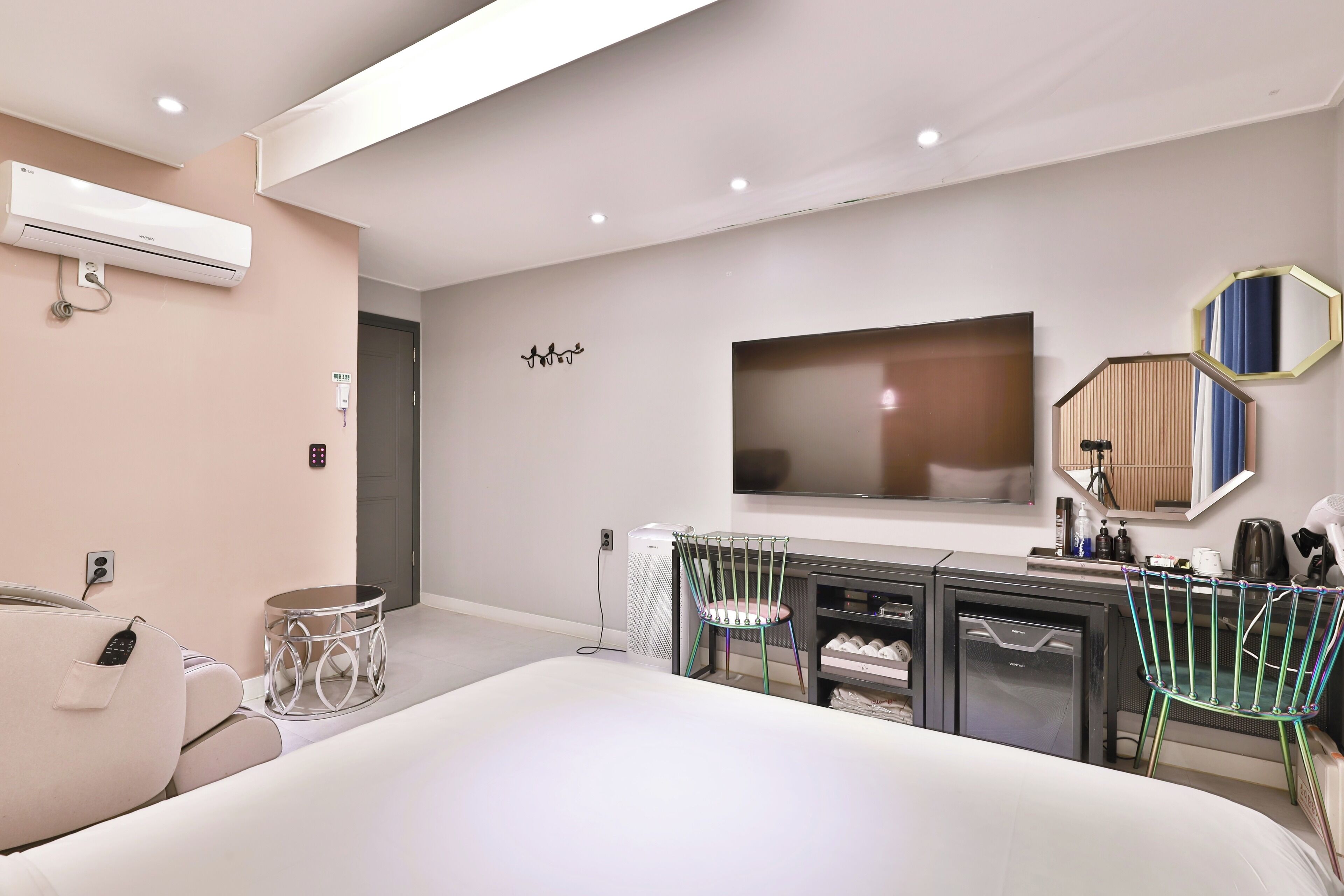 Una habitación de hotel moderna con una cama grande, un televisor montado en la pared, una kitchenette pequeña y una zona de estar con una silla y una mesita.