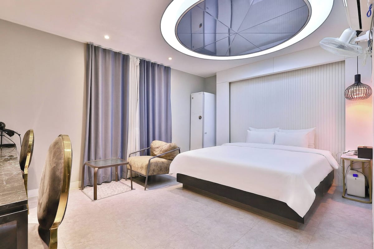A modern hotel room with a large bed, a desk with a chair, a small table, and a ceiling light fixture.