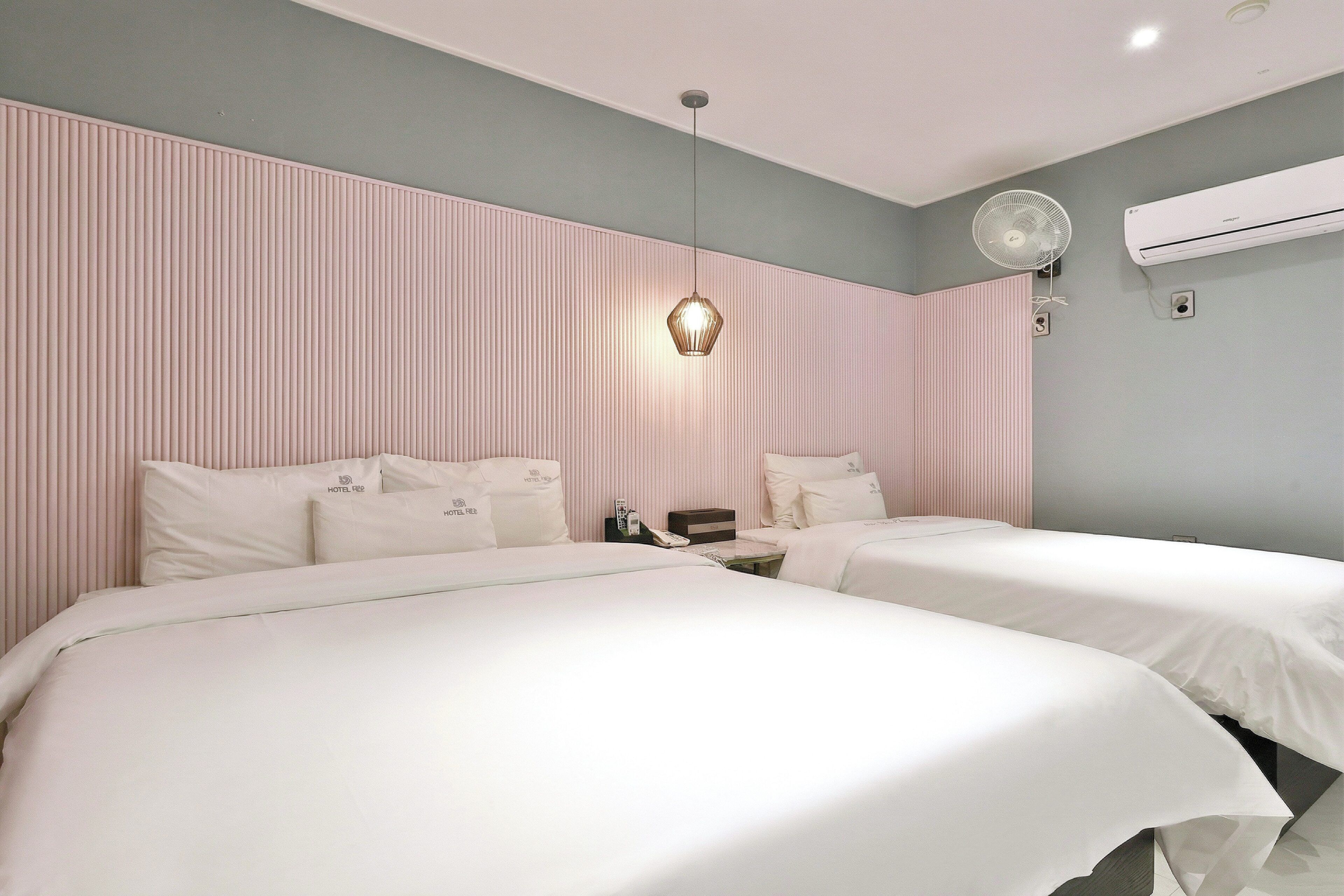 A hotel room with two beds, a hanging light fixture, an air conditioner, and a wall-mounted fan.