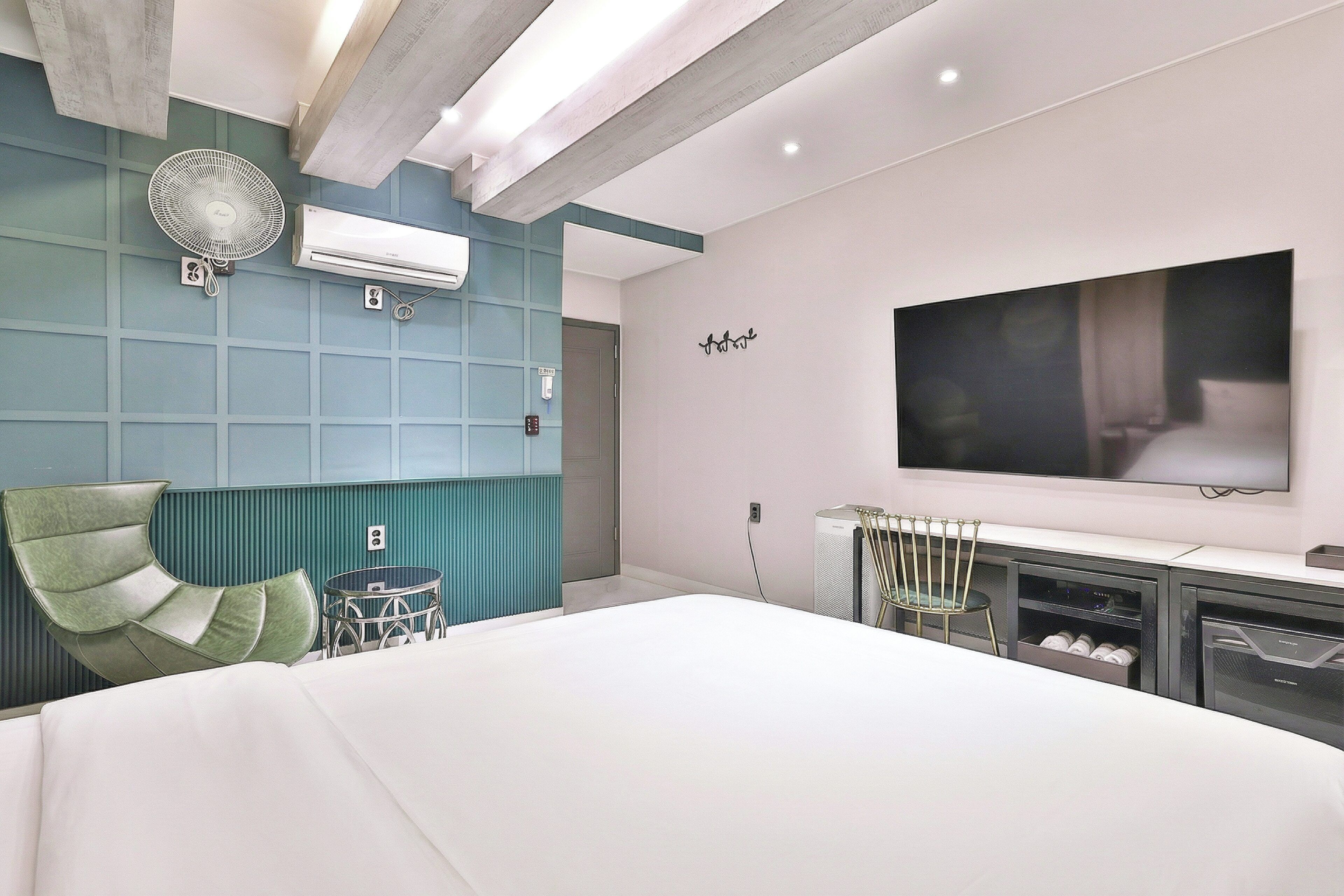 A modern hotel room with a large bed, a flat-screen TV mounted on the wall, a standing fan, and a small table with a chair.