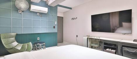 A modern hotel room with a large bed, a flat-screen TV mounted on the wall, a standing fan, and a small table with a chair.