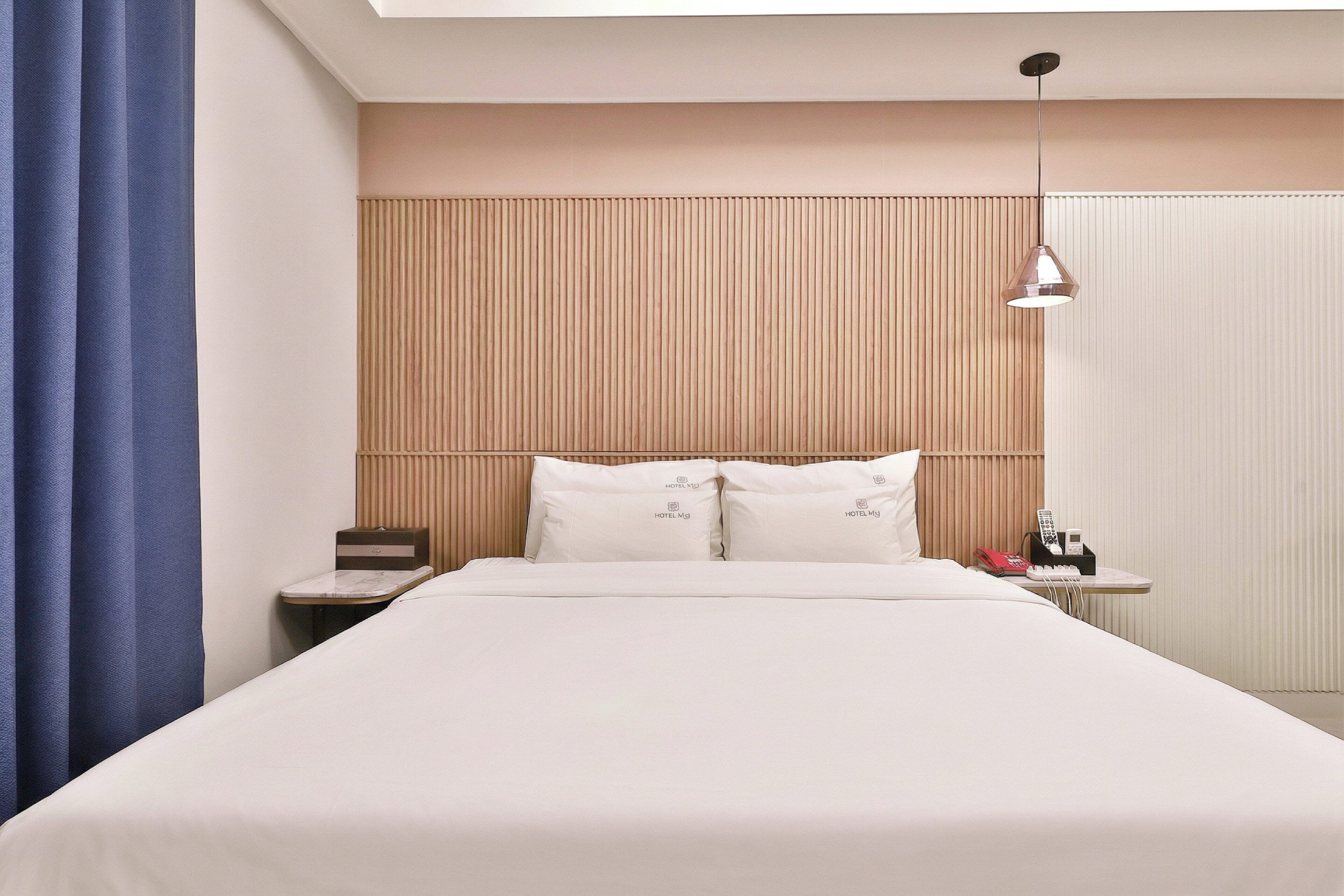 A hotel room with a large bed, a bedside table, a lamp, and a window with blinds.