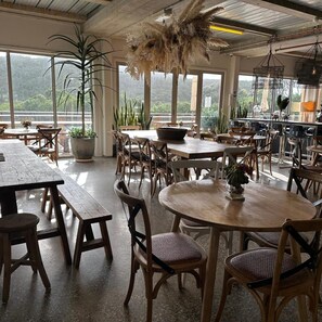 A spacious dining area with wooden tables and chairs, large windows offering views of greenery, and hanging plants. - Port Campbell Hostel (Port Campbell)