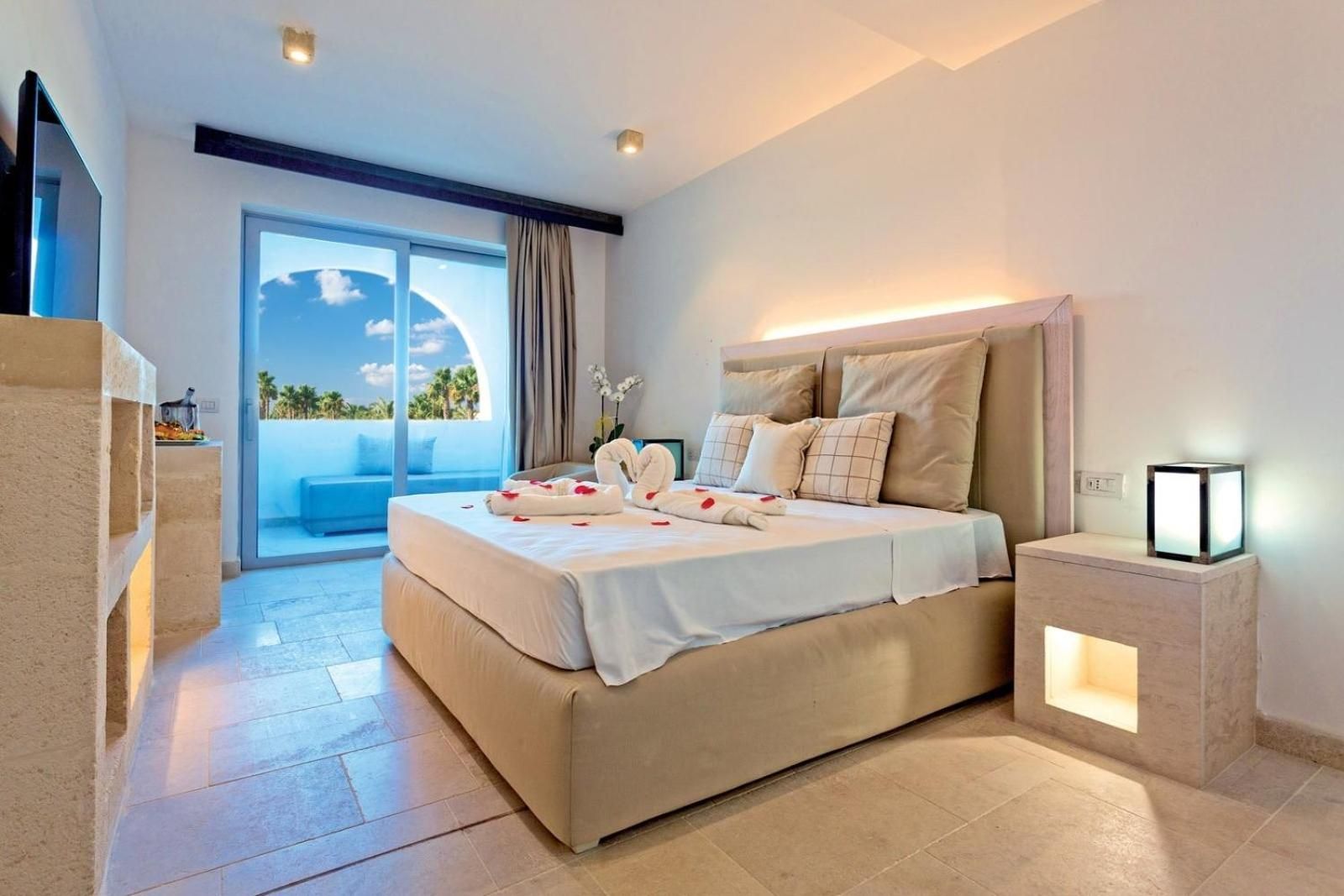 A modern hotel room with a large bed, a bedside table with a lamp, and a view of the outdoors.
