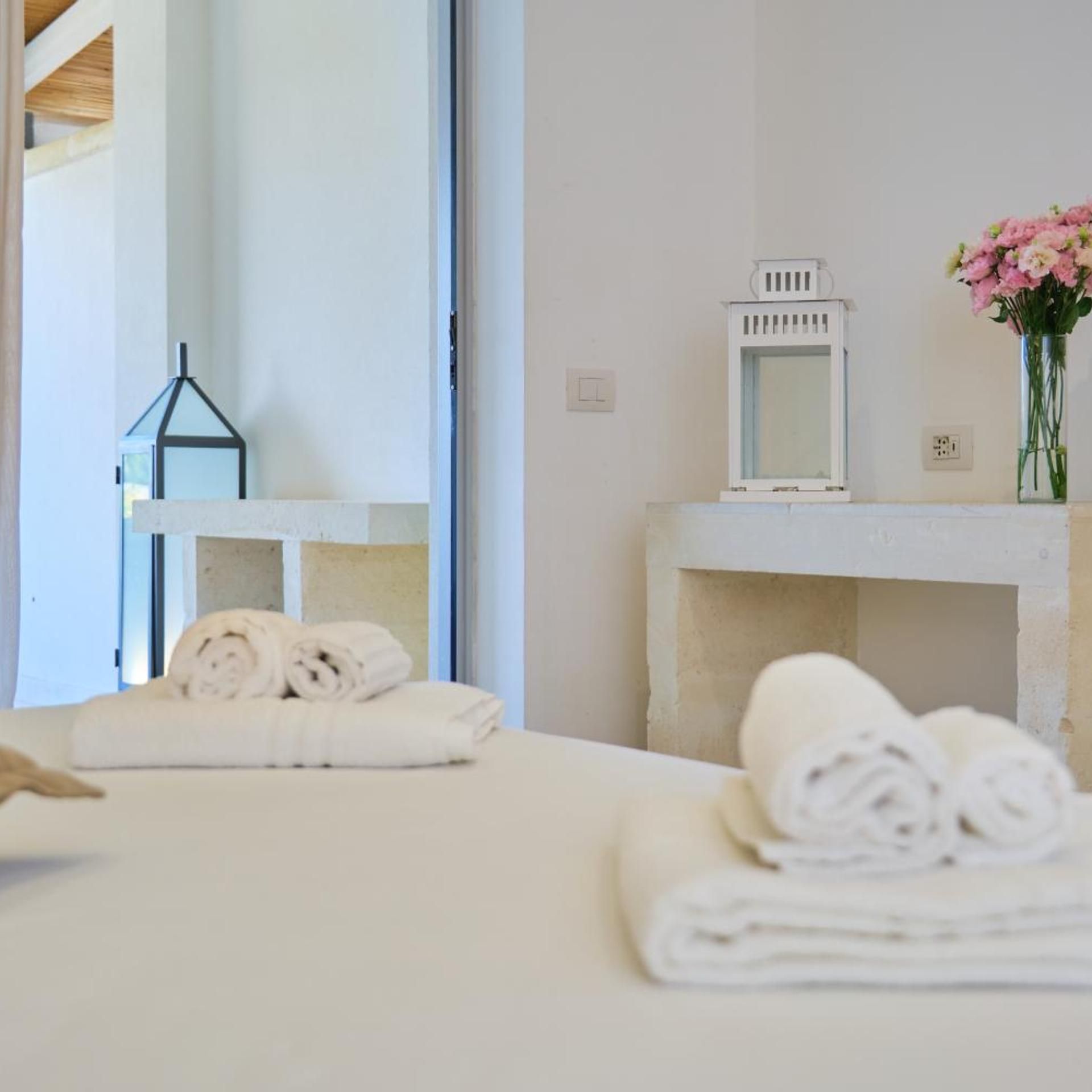 A hotel room with a bed, a bedside table with a mirror, a vase with flowers, and a towel rack.