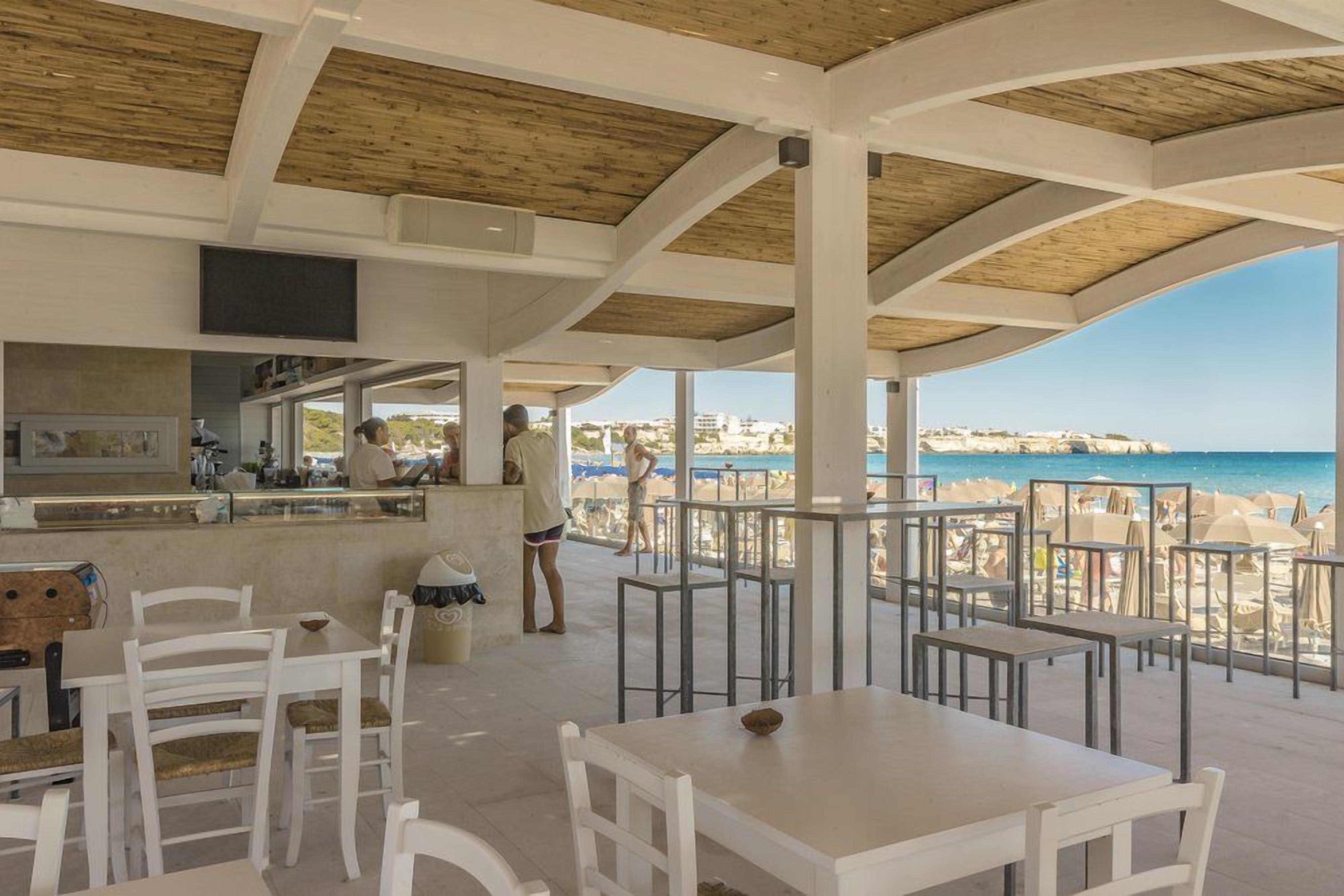 Area esterna con zona pranzo, tavoli e sedie bianche, un bancone bar e vista sulla spiaggia e sul mare.