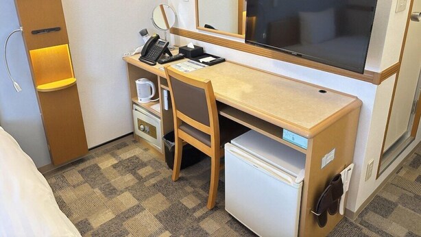 A hotel room with a desk, chair, refrigerator, microwave, coffee maker, telephone, mirror, and a large TV.