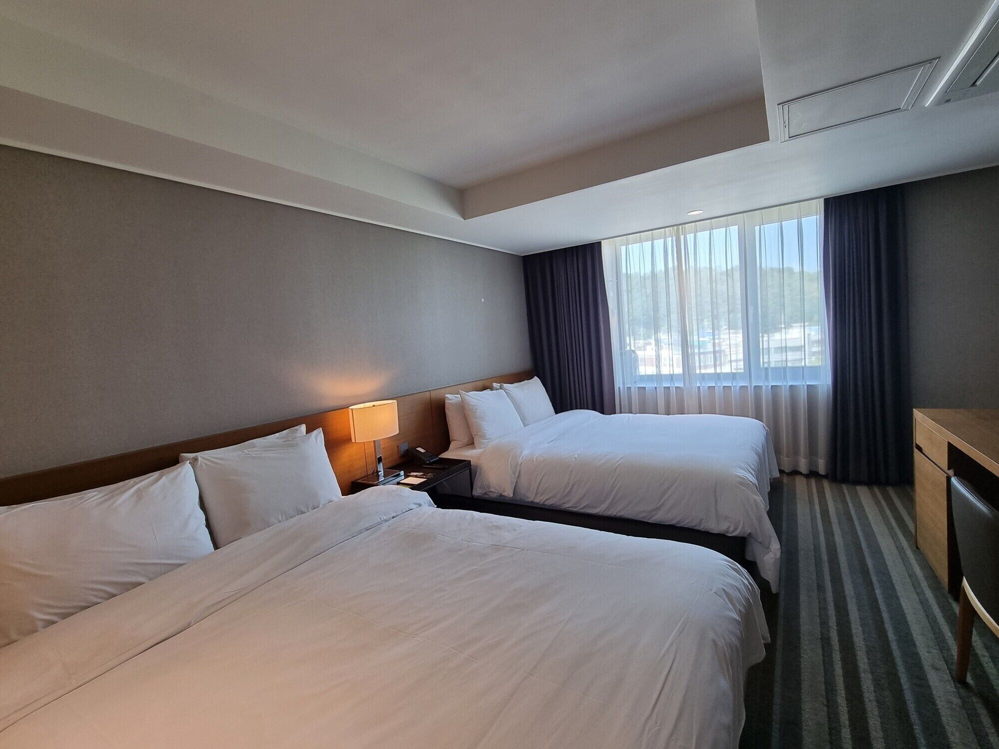 A hotel room with two beds, a desk, a lamp, and a window with curtains.