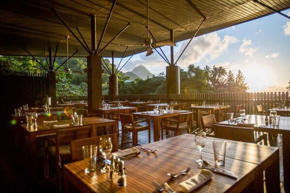 An outdoor restaurant with wooden tables and chairs, set against a backdrop of lush greenery and a mountain. - Boucan By Chocolat Hotel (Soufrière)