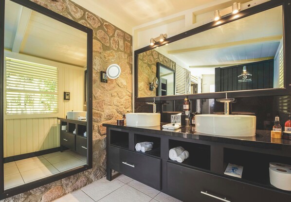 A bathroom with a stone wall, a large mirror, two sinks, and a vanity with drawers. - Boucan By Chocolat Hotel (Soufrière)