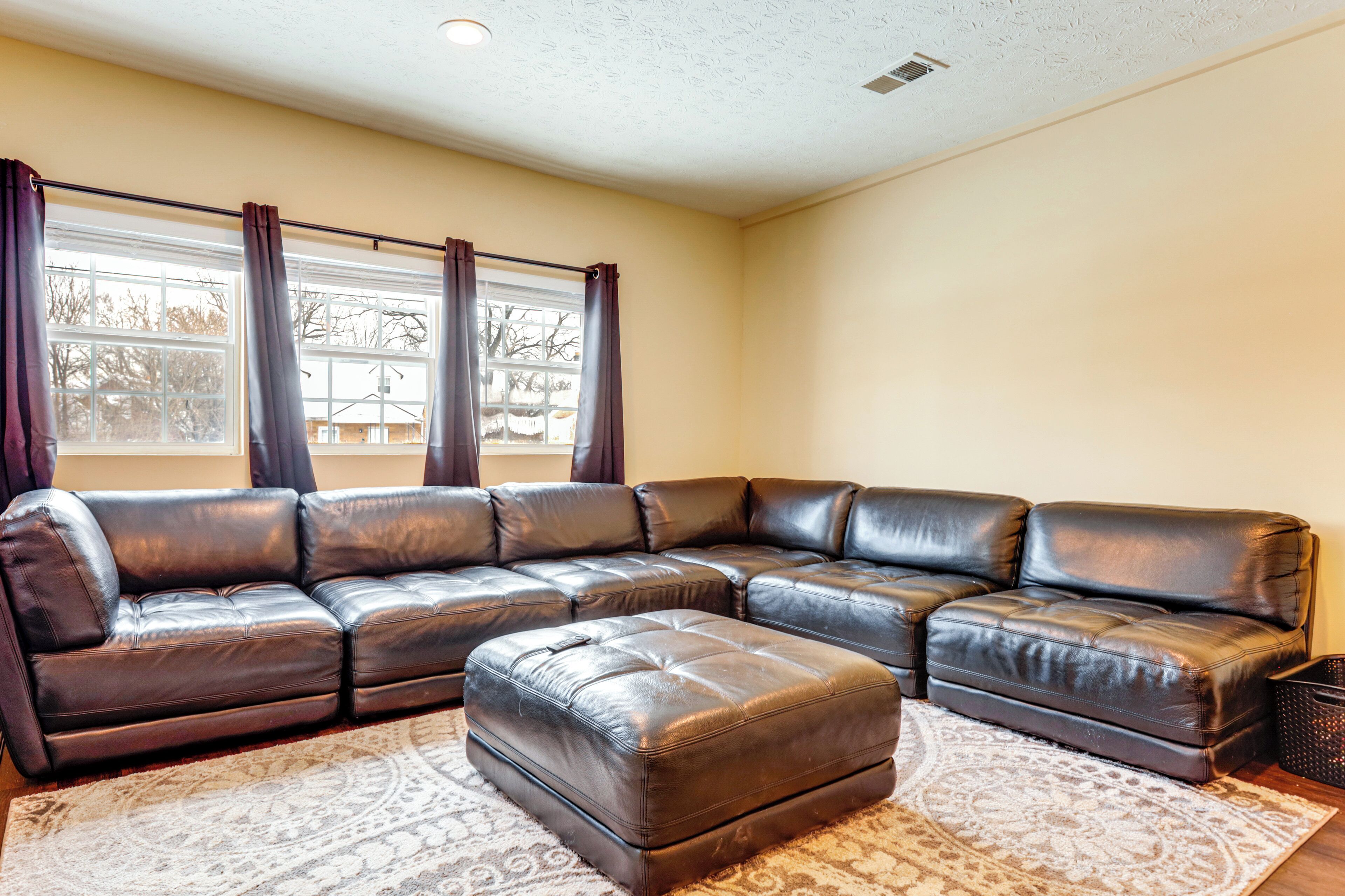 A sectional sofa and ottoman in a living room with a patterned rug, a window with curtains, and a ceiling with a recessed light.