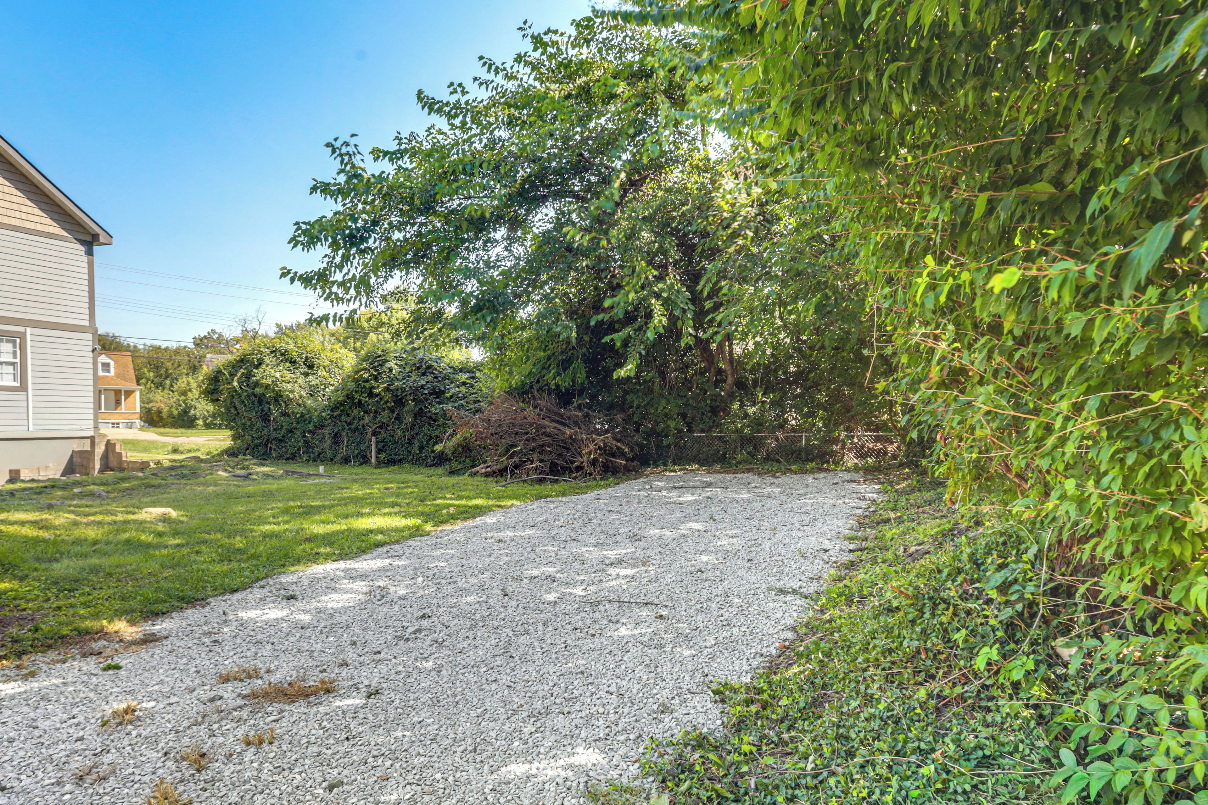 A gravel path leading to a house with a covered porch, surrounded by dense greenery and a clear sky.