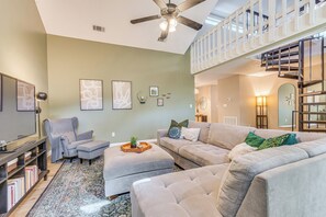 A spacious living room with a high ceiling, a ceiling fan, a large grey sofa, a grey armchair, a coffee table with a tray, and a staircase leading to an upper level. - Cottage Vibes in Shalimar! Pet-friendly Townhome (Shalimar)