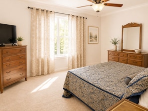 Quarto com cama, penteadeira, televisão, ventoinha no teto e janela com cortinas.