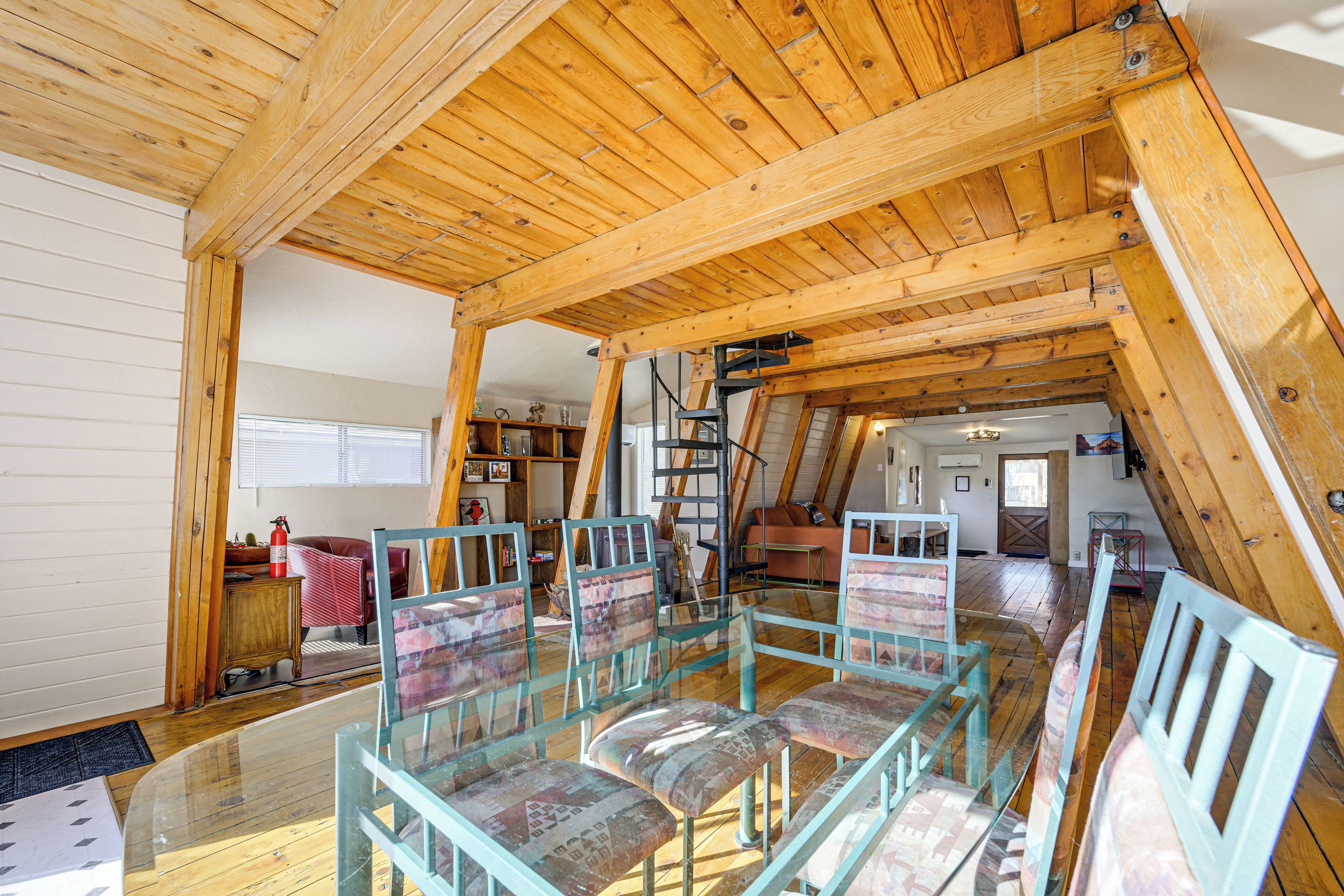 Amplia habitación en ático con mesa y sillas de vidrio, vigas de madera y zona de descanso elevada.