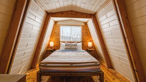 A narrow attic bedroom with wooden walls, a sloped ceiling, a bed with a striped mattress, two bedside lamps, and a window with blinds. - Hike, Bike & Atv! A-frame Rimrock Cabin w/ Views (Rimrock)