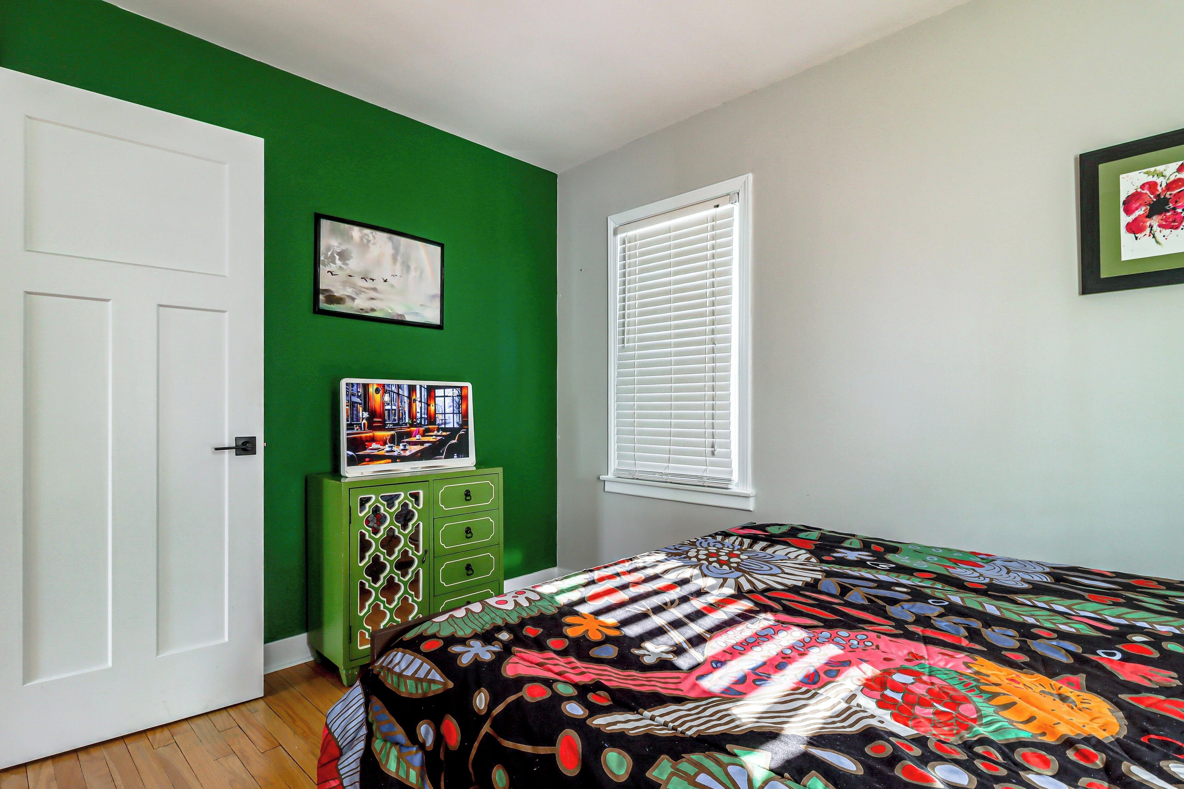 A bedroom with a green wall, a white door, a window with blinds, a bed with a colorful quilt, and a small cabinet with a picture on top.