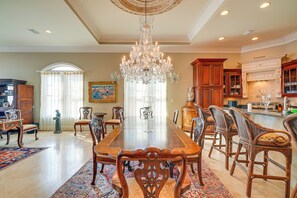 Uma ampla sala de jantar com uma grande mesa de madeira, cadeiras elaboradas, um lustre e uma cozinha ao fundo.