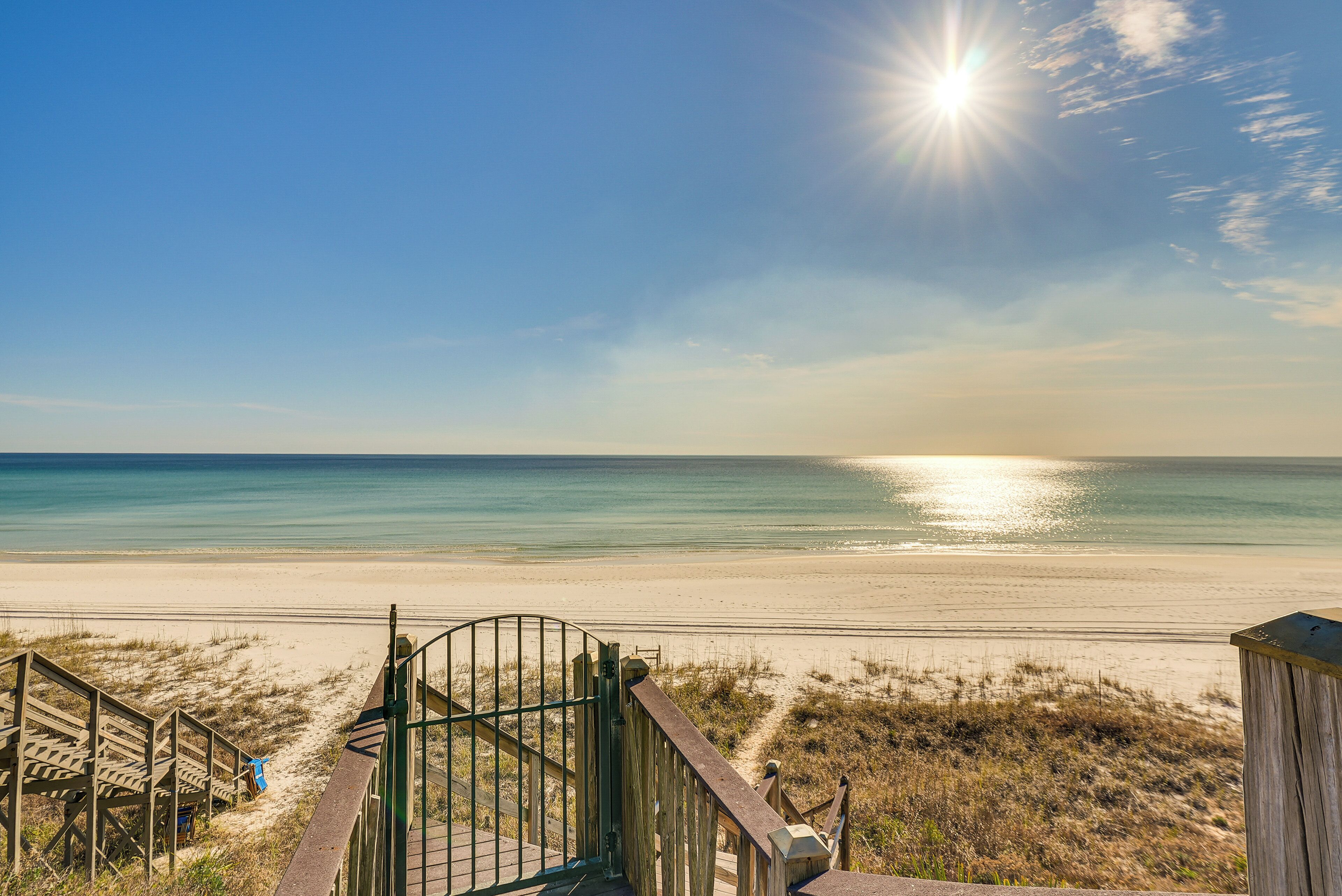 Ein hölzerner Steg führt zu einem Strand mit freiem Blick auf das Meer und einem sonnigen Himmel.