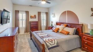 Quarto com cama, penteadeira de madeira, ventoinha no teto e janela com cortinas.