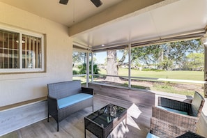 Un patio cubierto con mobiliario de mimbre, una mesa de vidrio y ventanas amplias con vistas a un campo de golf.