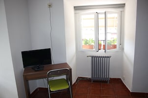 Quarto pequeno com escrivaninha, cadeira, radiador e janela com vista para plantas em vasos lá fora.