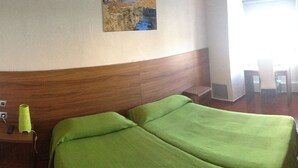 Una habitación de hotel con cabecera de madera, cama con ropa de cama verde, mesita de noche con lámpara y televisor montado en la pared.