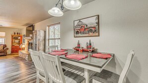 Un comedor con una mesa de madera, sillas blancas y manteles individuales rojos. En la pared, un cuadro de un camión rojo y una ventana con persianas.