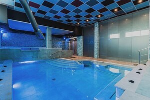 Una piscina interior moderna con paredes de vidrio transparente, piso de azulejos azules y techo a cuadros.