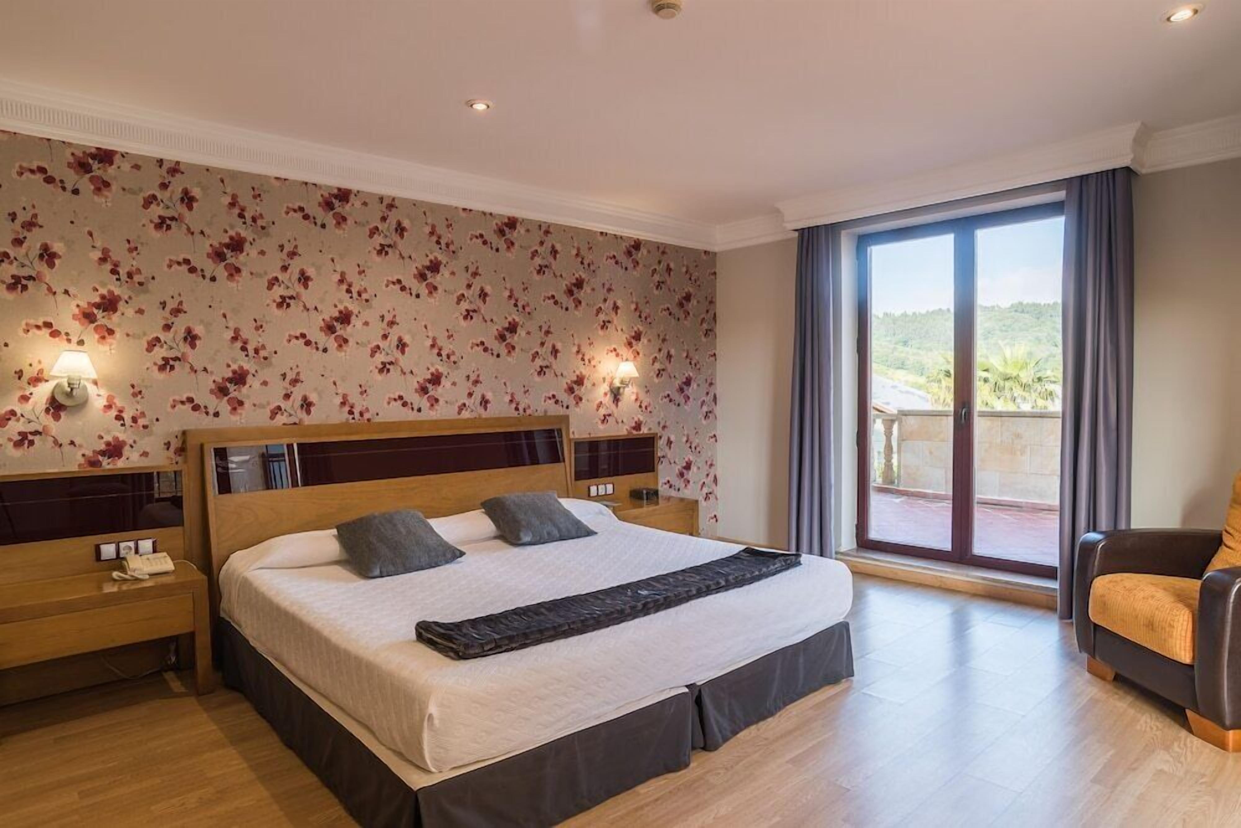 A hotel room with a large bed, a nightstand, a chair, and a balcony with a view.