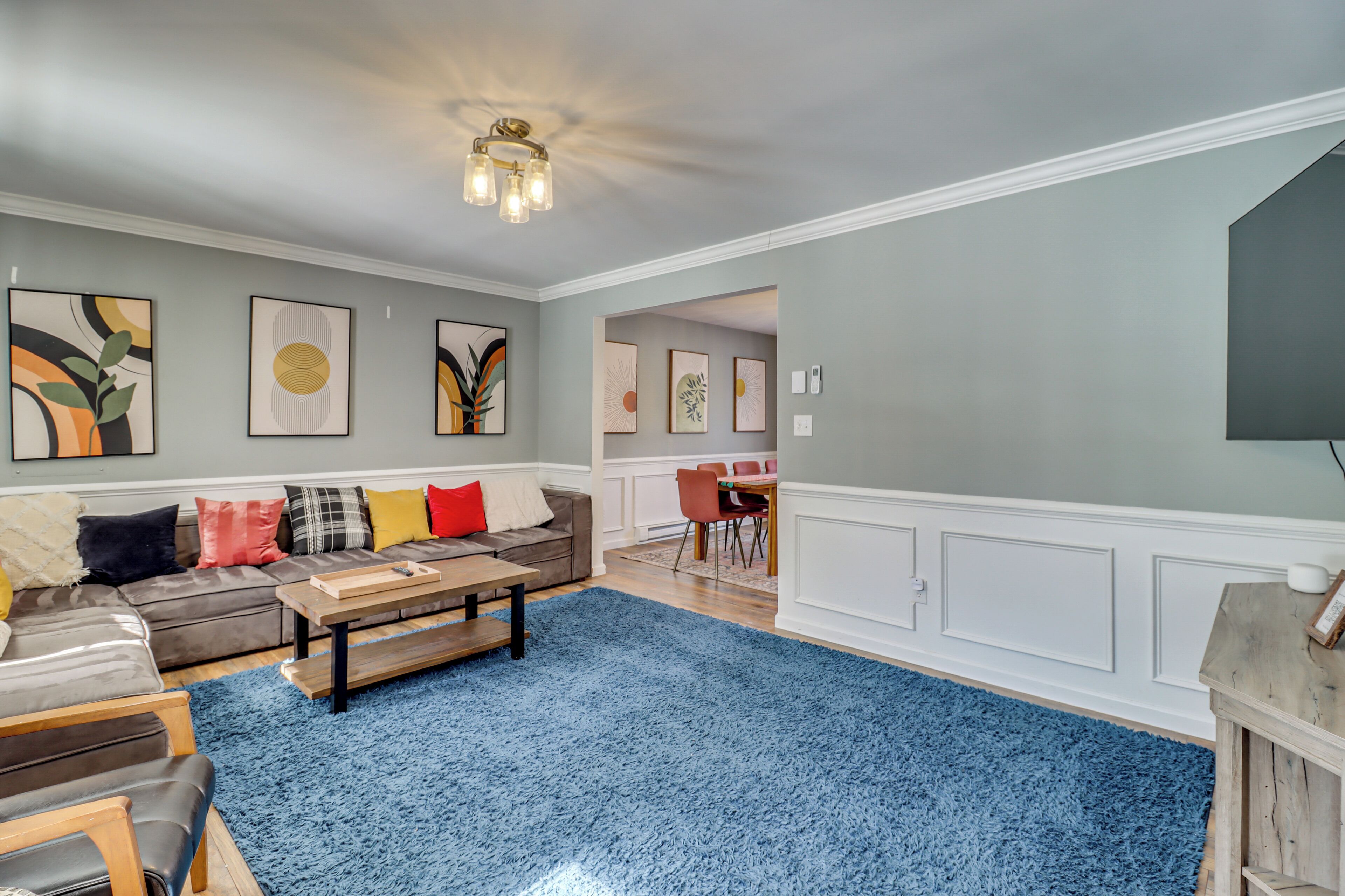 A living room with a blue carpet, a wooden coffee table, a grey sofa with colorful cushions, and framed artwork on the walls.