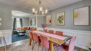 A dining area with a wooden table, pink chairs, and a chandelier. There are framed artworks on the walls and a blue rug on the floor.