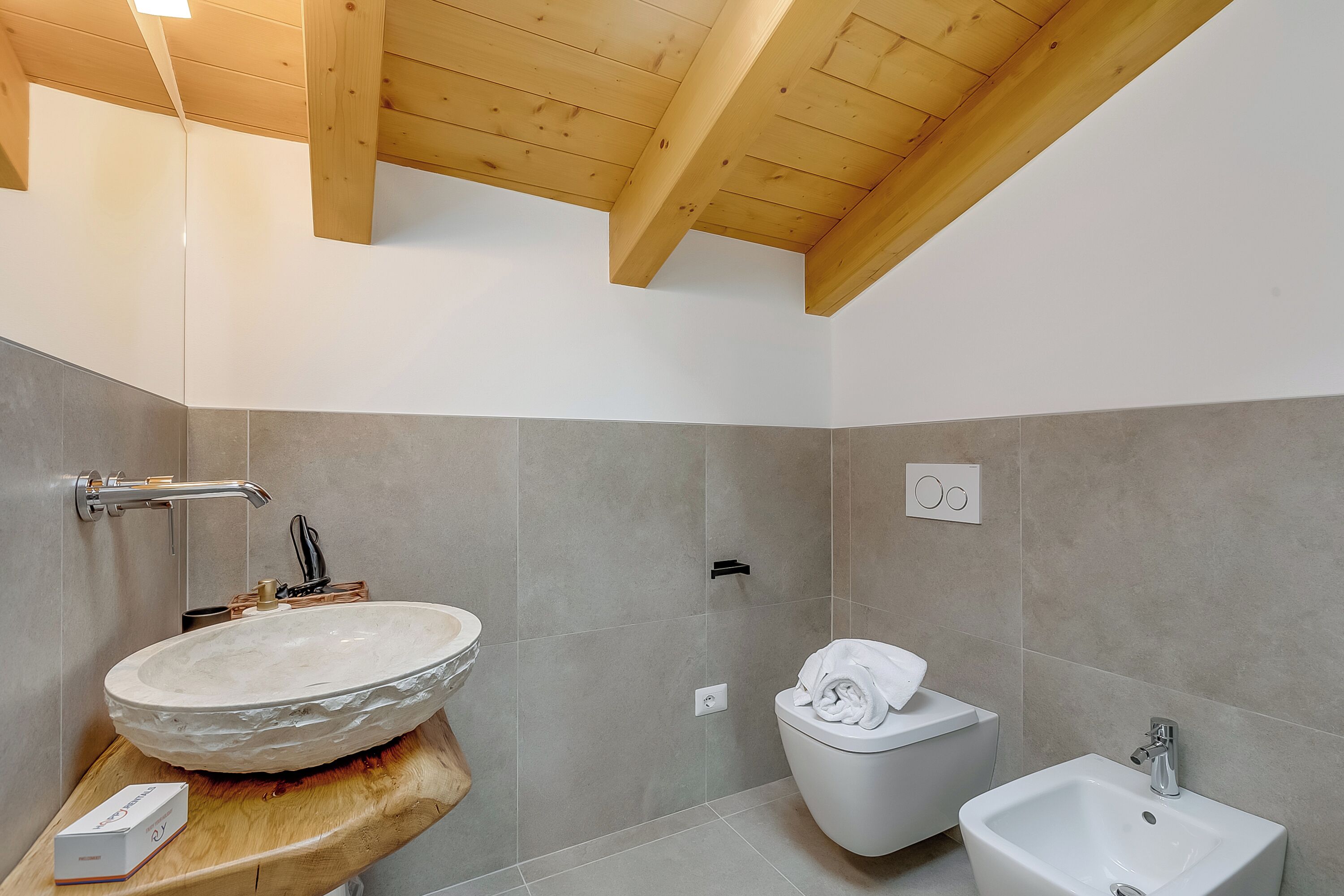 A modern bathroom with a stone sink, toilet, and bidet, wooden ceiling beams, and tiled walls.