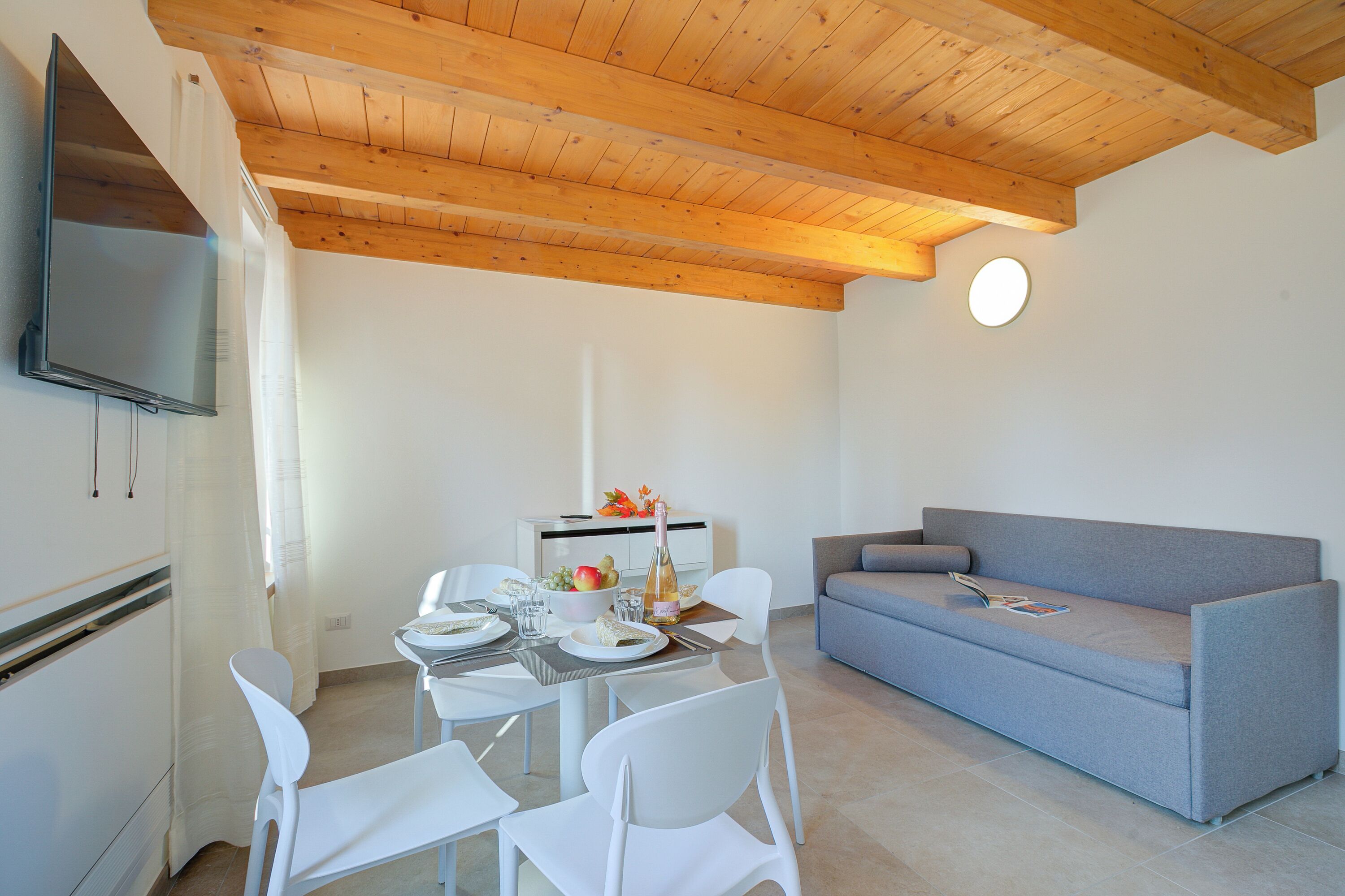 Un salon moderne comprenant un coin repas, des poutres en bois au plafond, un canapé gris et un téléviseur à écran plat fixé au mur.