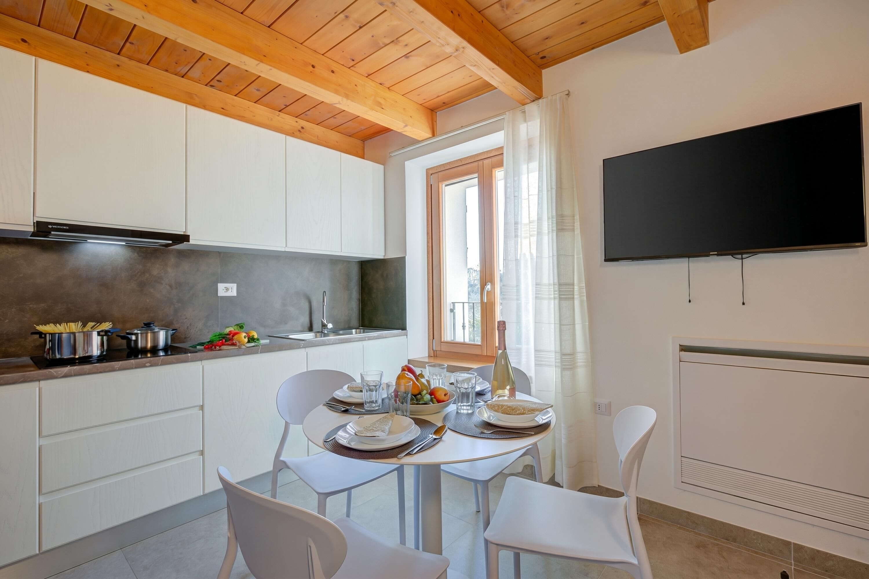 A modern kitchen with white cabinets, a dining table set with plates, glasses, and a bottle, and a flat-screen TV mounted on the wall.