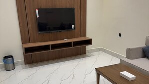 Ένα μοντέρνο σαλόνι με μια τηλεόραση επίπεδης οθόνης τοποθετημένη πάνω σε ένα ξύλινο έπιπλο, ένα δάπεδο με σχέδιο μαρμάρου και έναν γκρι καναπέ.