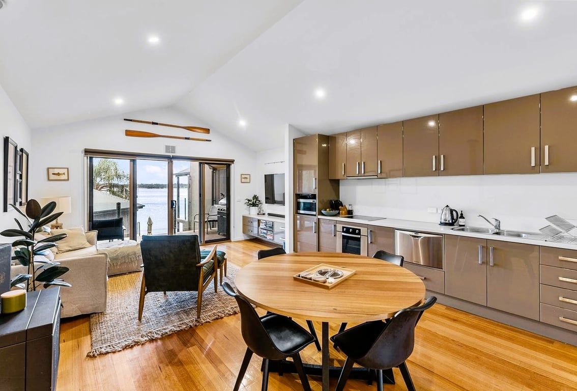 A modern kitchen with stainless steel appliances, a dining area with a round wooden table and black chairs, and a view of the outdoors through a glass door.