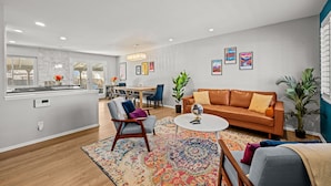 Ein modernes Wohnzimmer mit einem braunen Ledersofa, einem runden Couchtisch und einem farbenfrohen Teppich. Es gibt eine Essecke mit Tisch und Stühlen, sowie eine Küchenzeile mit Fenster und Pflanze.