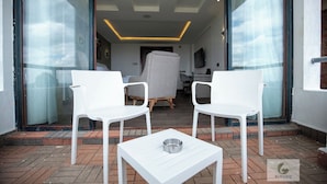 Un área de estar moderna al aire libre con sillas blancas y una mesa, una entrada con puerta de cristal y un espacio interior visible con una luz de techo.