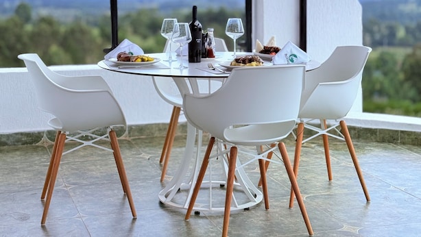 Mesa para dos con sillas blancas, una botella, copas de vino y platos de comida en un balcón con vista al paisaje.