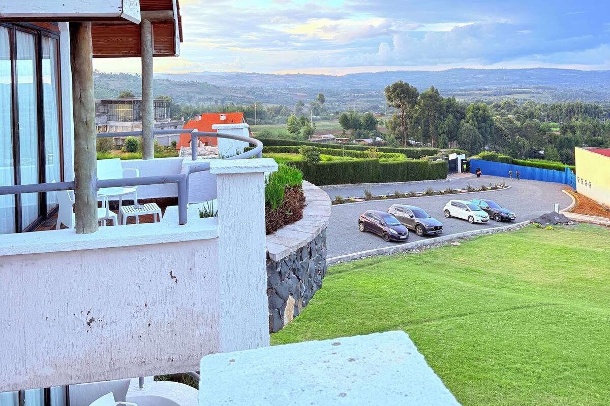 A building with a red roof, a balcony with white chairs and tables, and a view of a parking area with cars and a green landscape.