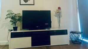 Ett vardagsrum med en platt-TV placerad på en vit TV-bänk, en krukväxt, en vas med blommor och två inramade konstverk på väggen.