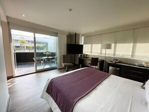 A modern hotel room with a large bed, a desk, a chair, a TV, and a sliding glass door to the outside. - BH Poblado (Medellín)