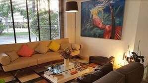 A living room with a beige sofa, a black leather couch, and a glass coffee table. There is a painting on the wall and a lamp on a side table. - La Sort Hotel (Teulada)