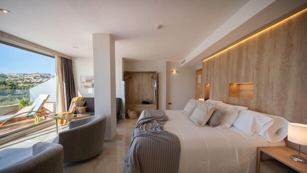 A modern hotel room with a large bed, a seating area with a sofa and armchairs, a balcony with a view of the hills, and a wooden accent wall. - La Sort Hotel (Teulada)