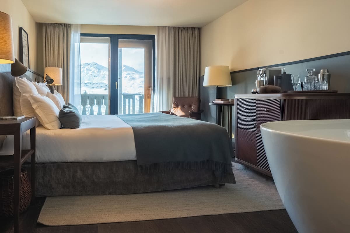 A hotel room with a bed, a sitting area with a chair, a large window with a view of mountains, a bathroom with a bathtub, and a minibar.