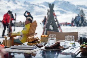 A table with food and drinks, a chair, and a view of a snowy mountain landscape. - Le Fitz Roy (Les Belleville)