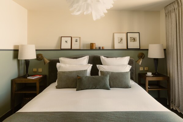 A hotel room with a large bed, two bedside tables with lamps, a wall-mounted shelf with framed pictures, and a hanging light fixture. - Le Fitz Roy (Les Belleville)